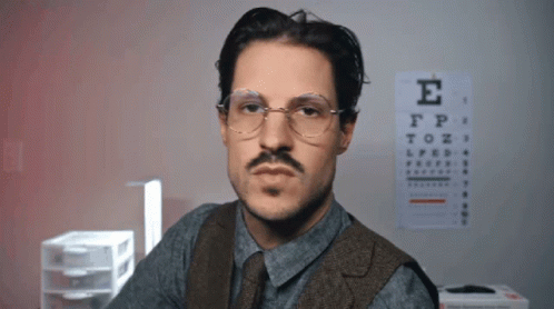 A Man With Glasses And A Mustache Looks At The Camera In Front Of An Eye Chart With The Letter E On It GIF