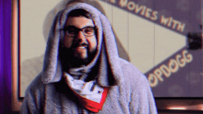 A Man With Glasses And A Scarf Around His Neck Stands In Front Of A Sign That Says Movies With Opdogg GIF