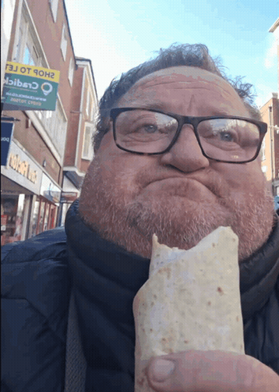A Man With Glasses Is Eating A Burrito In Front Of A Sign That Says ' Shop Ot 9Oh ' On It GIF