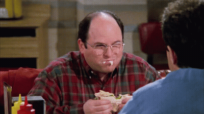 A Man With Glasses Is Eating A Sandwich While Another Man Watches GIF
