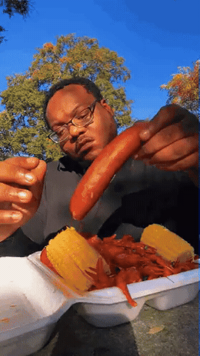 A Man With Glasses Is Eating Corn On The Cob And Crayfish GIF