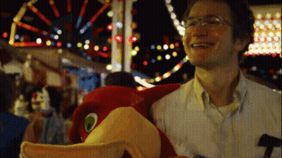 A Man With Glasses Is Holding A Stuffed Bird At A Carnival GIF