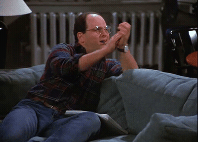 A Man With Glasses Is Sitting On A Couch With His Hands In The Air GIF