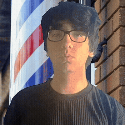 A Man With Glasses Stands In Front Of A Barber Pole GIF
