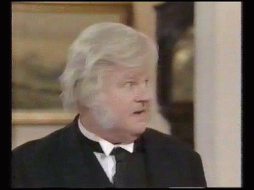 A Man With Gray Hair And A Beard Is Wearing A Black Suit And Tie . GIF
