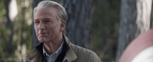 A Man With Gray Hair And A Brown Jacket Is Standing In Front Of A Tree And Looking At The Camera . GIF