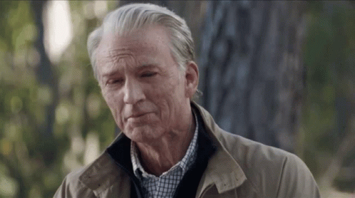A Man With Gray Hair And A Brown Jacket Is Standing In The Woods . GIF