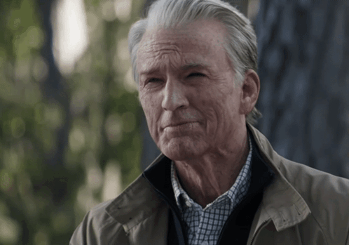 A Man With Gray Hair And A Tan Jacket Looks At The Camera GIF