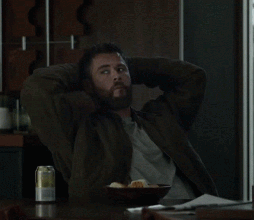A Man With His Hands Behind His Head Sits At A Table With A Bowl Of Food And A Can Of Beer GIF