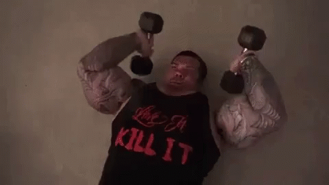 A Man With Huge Muscles Is Lifting Dumbbells While Wearing A Kill It Shirt . GIF