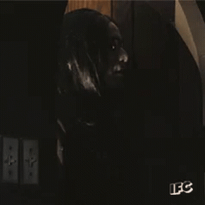 A Man With Long Black Hair And Glasses Is Standing In A Dark Room With A Sign That Says Ifc GIF