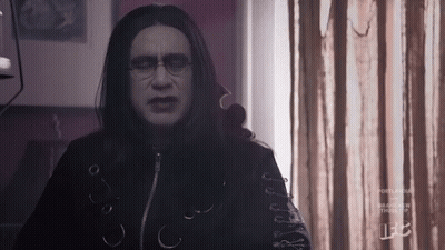 A Man With Long Black Hair And Glasses Is Standing In Front Of A Window In A Room . GIF