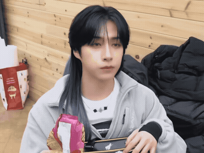 A Man With Long Black Hair Is Holding A Bag Of Chips In Front Of A Bag That Says Crayon Shin Chan GIF