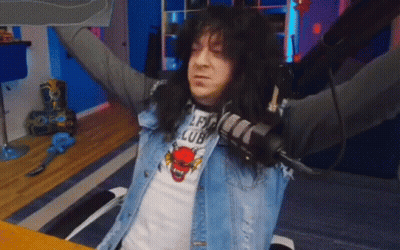 A Man With Long Black Hair Is Sitting At A Table With His Arms Outstretched And Holding A Microphone . GIF