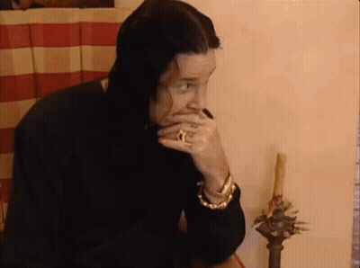 A Man With Long Black Hair Is Sitting At A Table With His Hand To His Mouth . GIF