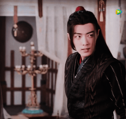 A Man With Long Black Hair Is Standing In A Room With A Candelabra In The Background GIF