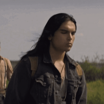 A Man With Long Black Hair Is Wearing A Denim Jacket And Standing In A Field . GIF