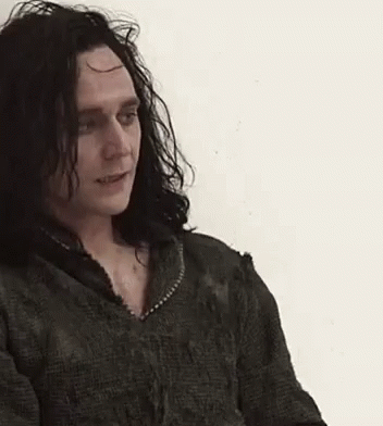 A Man With Long Black Hair Is Wearing A Green Sweater And Looking At The Camera . GIF