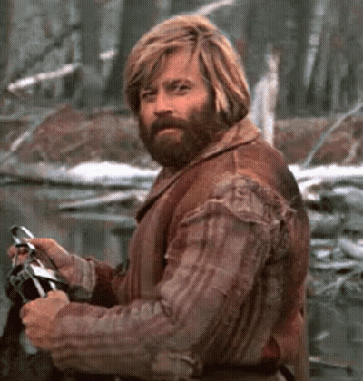 A Man With Long Blonde Hair And A Beard Is Holding A Fishing Rod In The Woods . GIF