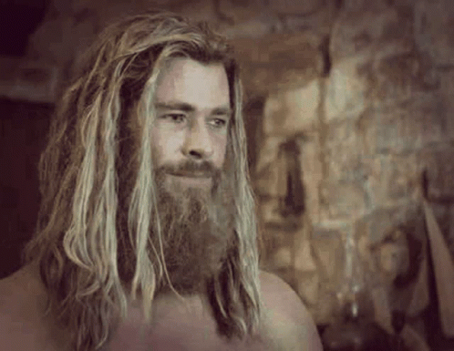 A Man With Long Blonde Hair And A Beard Is Standing In Front Of A Stone Wall . GIF
