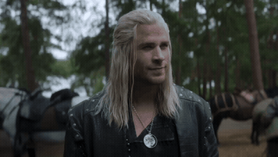 A Man With Long Blonde Hair And A Necklace Is Standing In The Woods . GIF