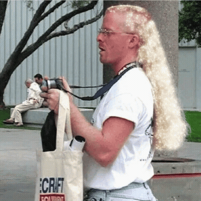 A Man With Long Blonde Hair Is Holding A Camera And A Tote Bag That Says Craft On It GIF