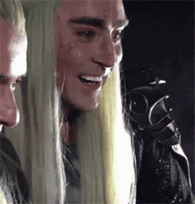 A Man With Long Blonde Hair Is Smiling And Looking At Another Man . GIF