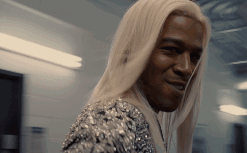 A Man With Long Blonde Hair Is Smiling And Wearing A Silver Dress GIF