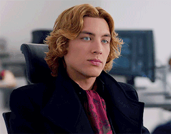 A Man With Long Blonde Hair Is Wearing A Black Coat And A Red Tie GIF
