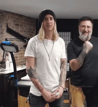 A Man With Long Blonde Hair Stands Next To Another Man With A Beard GIF