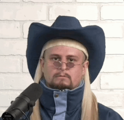A Man With Long Blonde Hair Wearing A Cowboy Hat And Sunglasses Is Standing In Front Of A Microphone . GIF