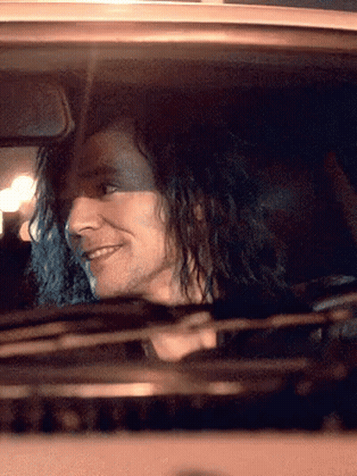 A Man With Long Curly Hair Is Driving A Car And Smiling GIF