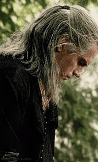 A Man With Long Gray Hair And A Black Shirt Is Standing In A Forest . GIF