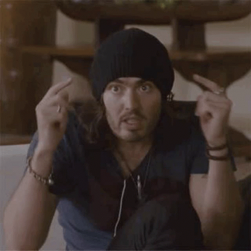 A Man With Long Hair And A Beanie Is Sitting On A Couch Making A Surprised Face . GIF
