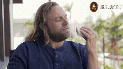 A Man With Long Hair And A Beard Is Holding A Napkin In Front Of A Dr. Squatch Logo GIF