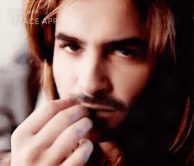 A Man With Long Hair And A Beard Is Holding His Hand To His Mouth . GIF