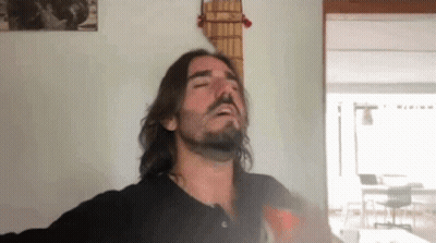 A Man With Long Hair And A Beard Is Smoking A Cigarette In Front Of A Guitar Hanging On A Wall . GIF