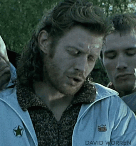 A Man With Long Hair And A Beard Is Smoking A Cigarette While Standing Next To Another Man . GIF