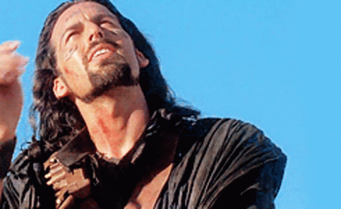 A Man With Long Hair And A Beard Is Standing In Front Of A Blue Sky Holding A Sword . GIF