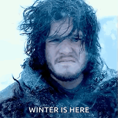 A Man With Long Hair And A Beard Is Standing In The Snow With The Words `` Winter Is Here '' Written Above Him . GIF