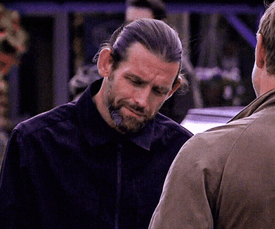 A Man With Long Hair And A Beard Is Talking To Another Man In A Brown Jacket GIF