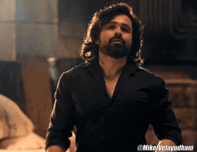 A Man With Long Hair And A Beard Is Wearing A Black Shirt With @Mikelvelayudham On The Bottom GIF