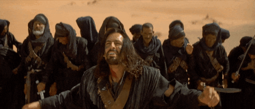 A Man With Long Hair And A Beard Stands In Front Of A Group Of People GIF