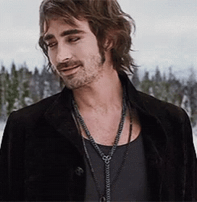 A Man With Long Hair And A Beard Wearing A Black Jacket And A Necklace . GIF