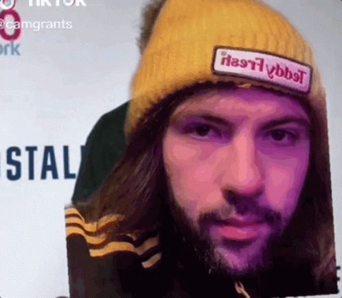A Man With Long Hair And A Beard Wearing A Yellow Beanie With A Patch That Says " Heavyybbt " GIF
