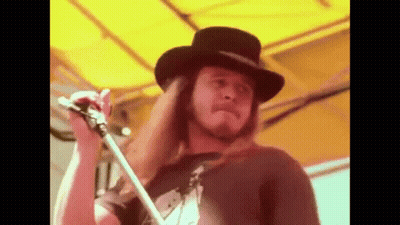 A Man With Long Hair And A Hat Is Holding A Microphone . GIF
