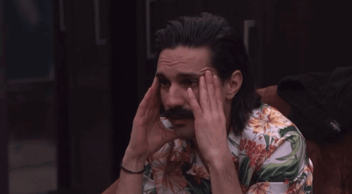 A Man With Long Hair And A Mustache Holds His Head GIF