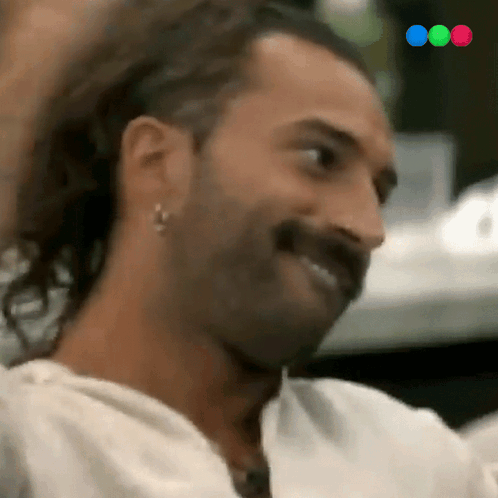 A Man With Long Hair And A Mustache Is Smiling While Wearing Earrings . GIF