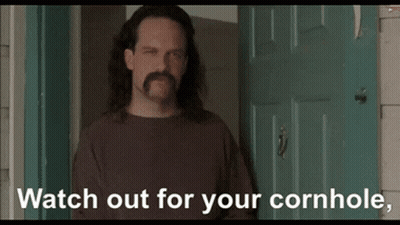 A Man With Long Hair And A Mustache Is Standing In Front Of A Door With The Words " Watch Out For Your Cornhole " Below  GIF