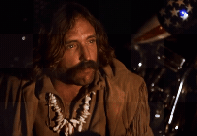 A Man With Long Hair And A Mustache Is Standing In Front Of A Motorcycle With An American Flag In The Background GIF
